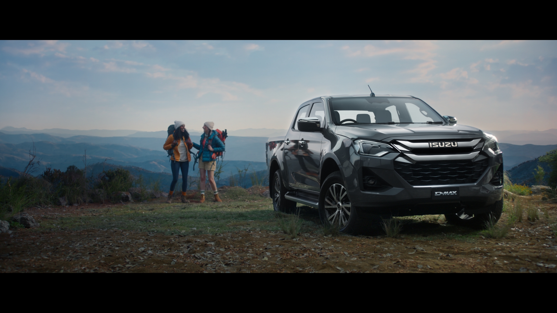 Isuzu D-Max parked near two hikers with backpacks and caps, mountain range in the background, clear sky, outdoor vehicle, auto advertisement.