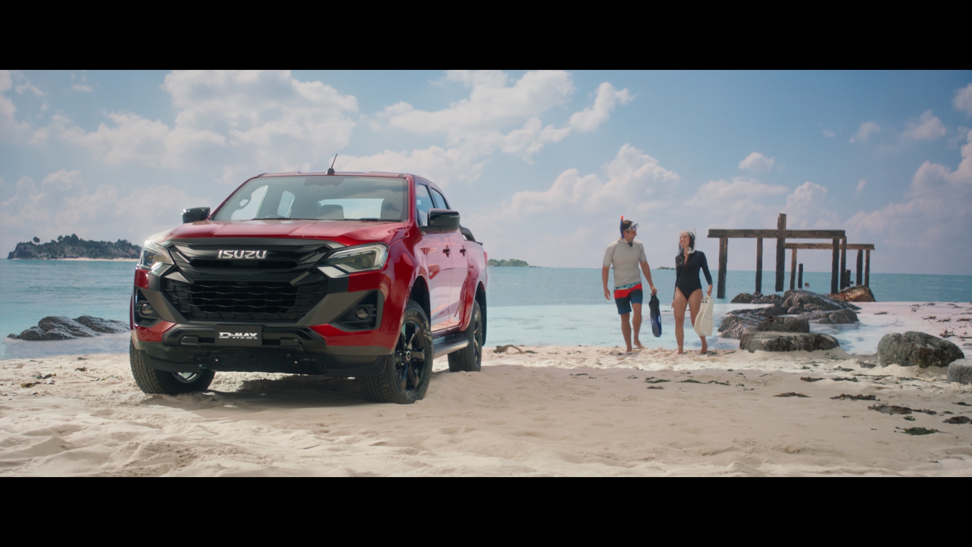 Red Isuzu D-Max pickup truck parked on a sandy beach with two snorkelers walking in the background on a sunny day near the ocean.