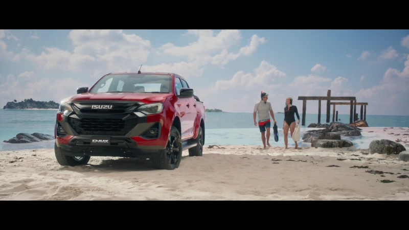 Red Isuzu D-Max pickup truck parked on a sandy beach with two snorkelers walking in the background on a sunny day near the ocean.