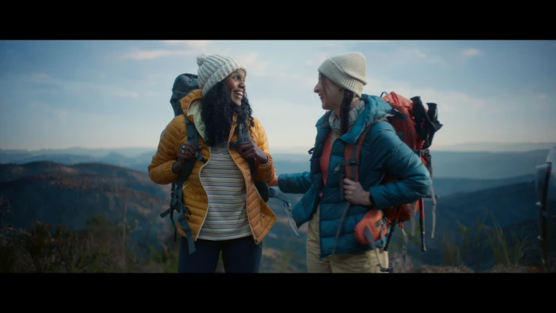 Two smiling women with backpacks wearing hats stand in a mountainous landscape under a blue sky with wispy clouds, enjoying outdoor activities.