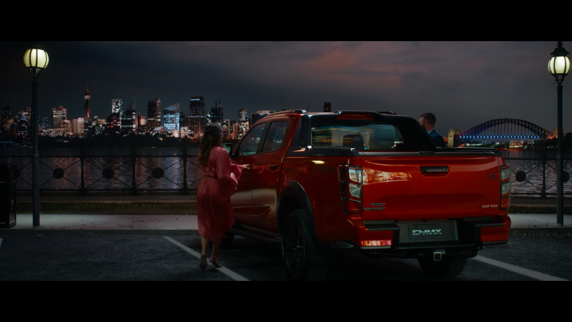 Red Isuzu D-Max parked in an urban setting at night, with a woman entering the vehicle and a man standing beside it, city skyline.
