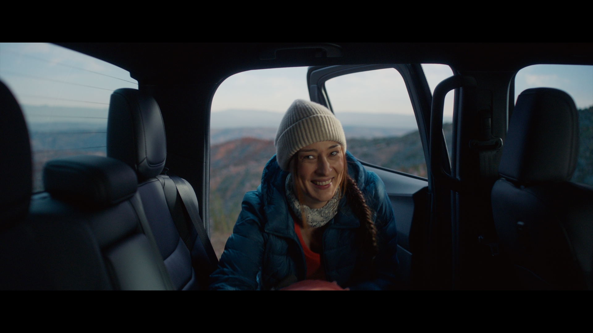 Woman with a beanie sits in the back of a car, smiling with mountain views in the distance during daytime, captured in a medium shot.