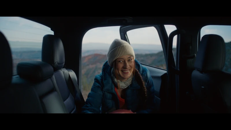 Woman with a beanie sits in the back of a car, smiling with mountain views in the distance during daytime, captured in a medium shot.