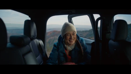 Smiling woman wearing a beanie and blue jacket sitting inside the back of a truck overlooking a mountainous landscape