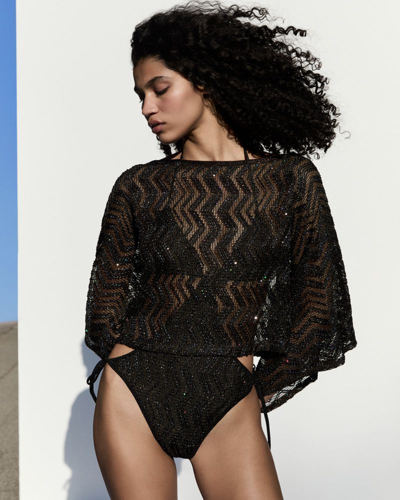 An adult female model poses in a black, shiny, zigzag-patterned swimwear set against a bright white backdrop, with natural outdoor light.