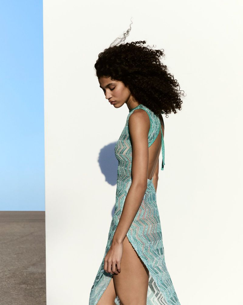 A woman with curly dark hair wearing a turquoise knitted dress stands against a white wall in an outdoor fashion shot.