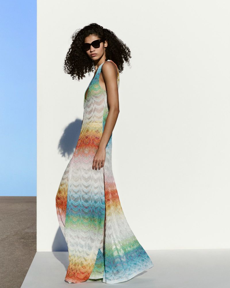 A woman in a rainbow striped dress and sunglasses poses in front of a white wall with a blue accent for a fashion editorial shot.