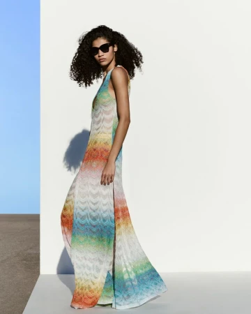 A woman in a rainbow striped dress and sunglasses poses in front of a white wall with a blue accent for a fashion editorial shot.
