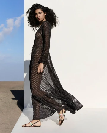 Model with curly dark hair wearing a long sheer black knit dress and strappy sandals outdoors against a white wall.