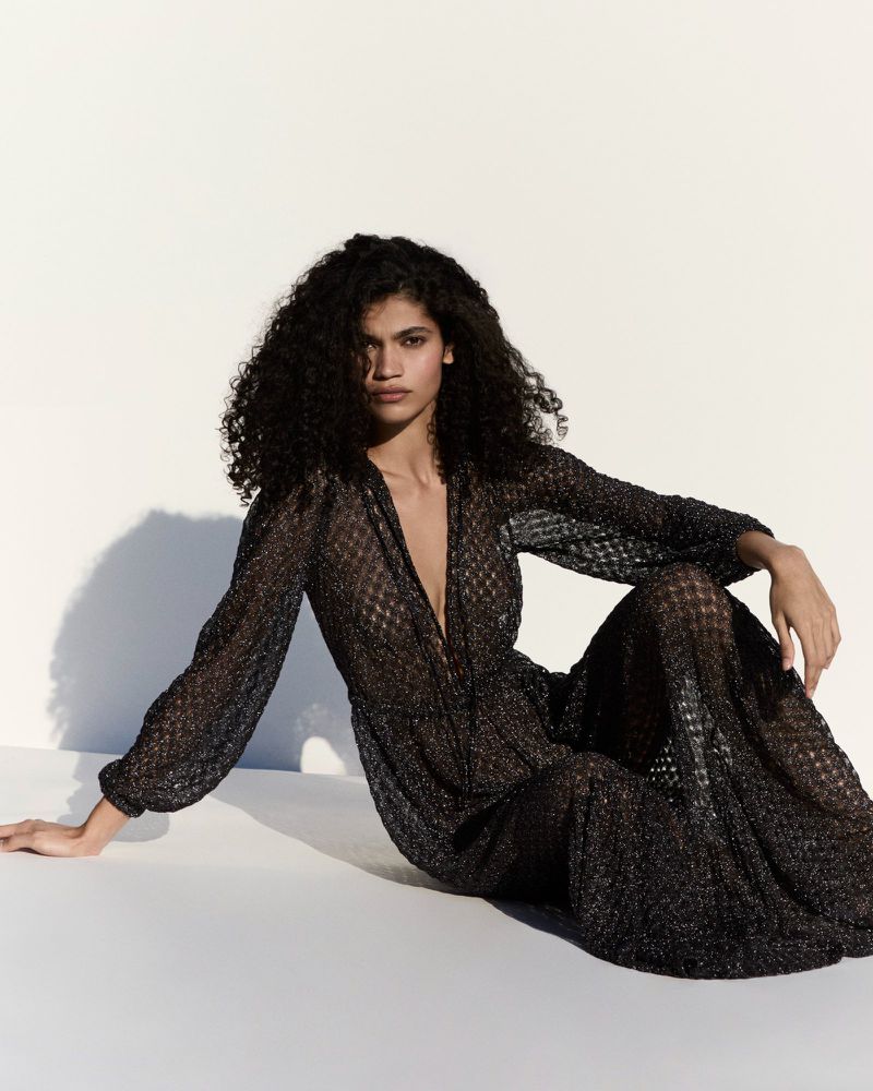 Model Raynara Negrine wearing a sheer patterned black maxi dress while seated against a bright white backdrop with cast shadows.
