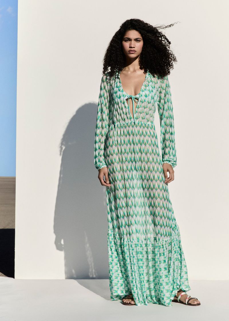 A model poses against a wall wearing a patterned, long-sleeved, ankle-length dress with sandals in an outdoor setting with blue sky.