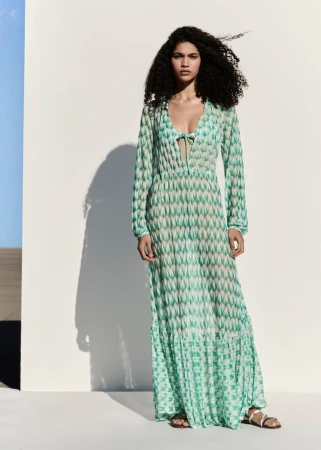 A model poses against a wall wearing a patterned, long-sleeved, ankle-length dress with sandals in an outdoor setting with blue sky.