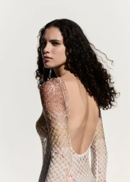 Model with long dark curly hair wearing a patterned open back dress against a clean white seamless studio background.
