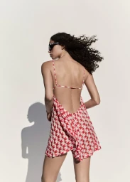 Model with dark curly hair wearing a patterned red and white backless dress against a plain white studio background.