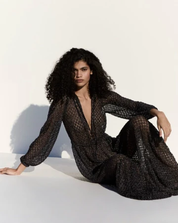 Model Raynara Negrine wearing a sheer patterned black maxi dress while seated against a bright white backdrop with cast shadows.