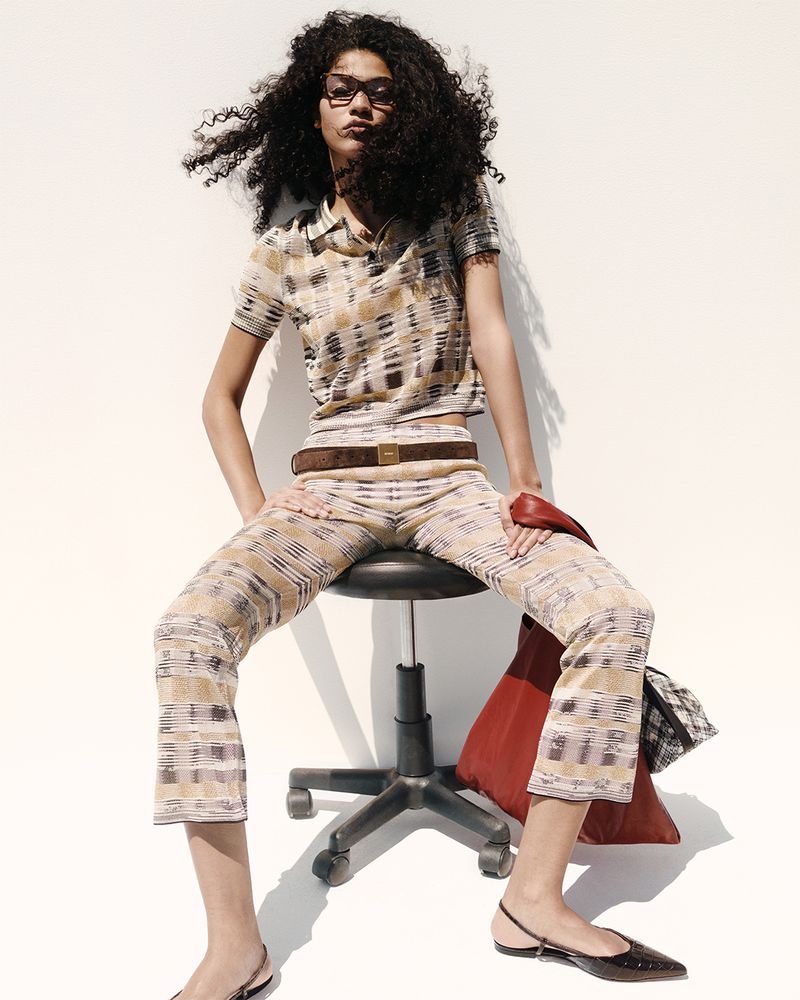 Woman sits on a stool in a patterned knit outfit with a brown belt, carrying a red leather tote bag. Studio shot against a white background.