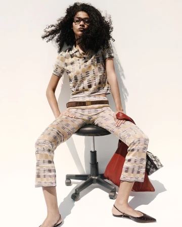 Woman sits on a stool in a patterned knit outfit with a brown belt, carrying a red leather tote bag. Studio shot against a white background.