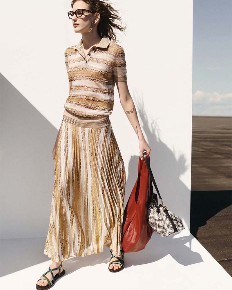 A woman in a tan knit outfit with a pleated skirt carries two bags against a split white and brown background on a sunny day.