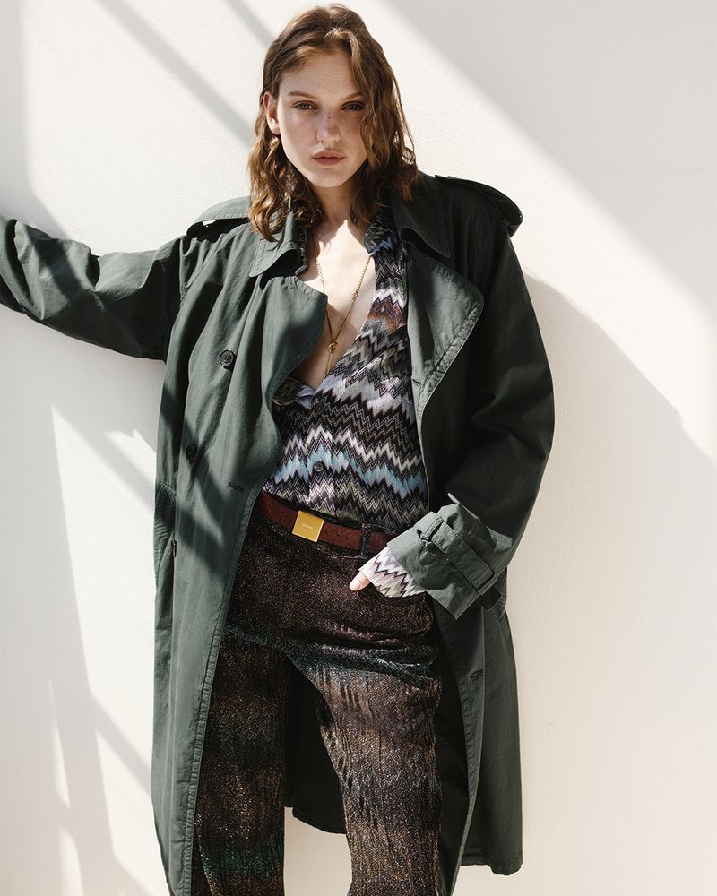Model posing in an oversized dark green trench coat and patterned Missoni chevron shirt against a sunlit white wall.