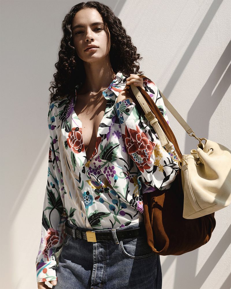 Woman in a floral blouse and jeans stands indoors with a brown and beige bucket bag on her shoulder, with natural, diffused light.