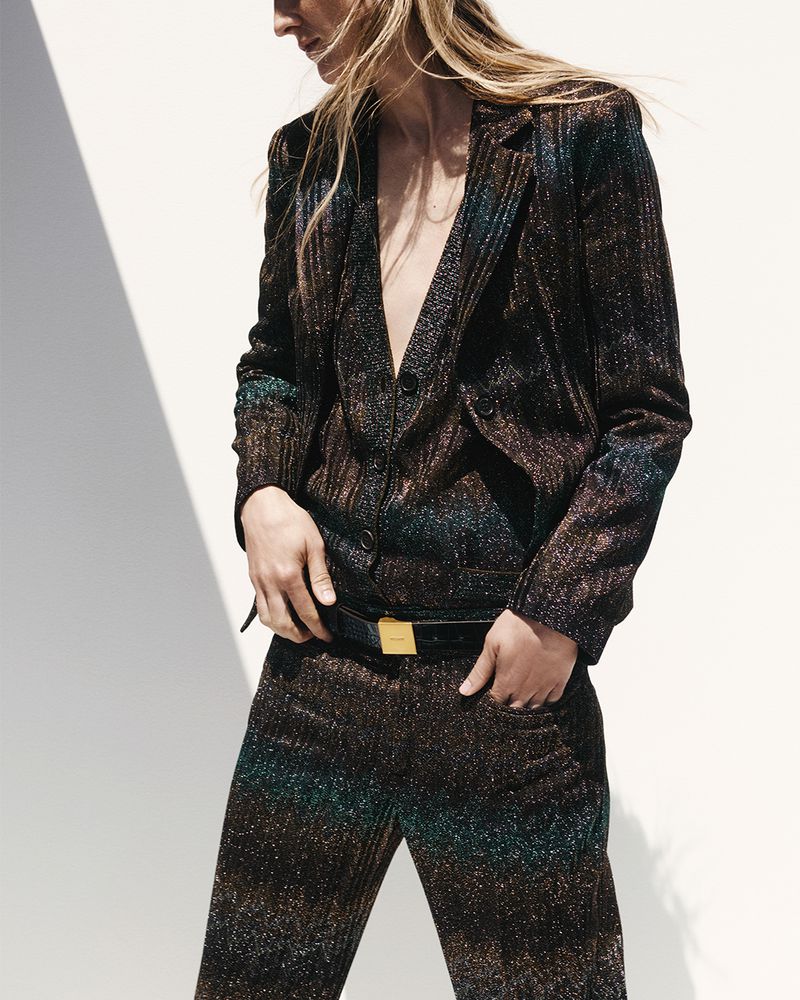 A woman poses in a sparkling, textured two-piece suit with a belt against a bright wall with sunlight casting shadows.
