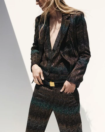 A woman poses in a sparkling, textured two-piece suit with a belt against a bright wall with sunlight casting shadows.
