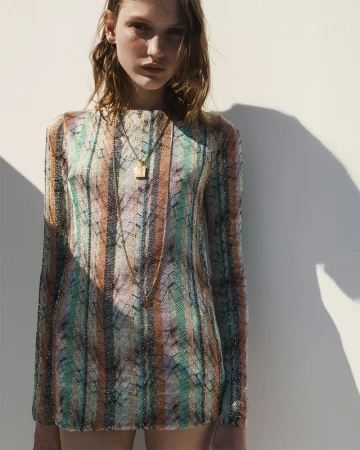 Model posing in a multicolored striped long-sleeve knit mini dress and gold pendant necklace against a sunlit wall.