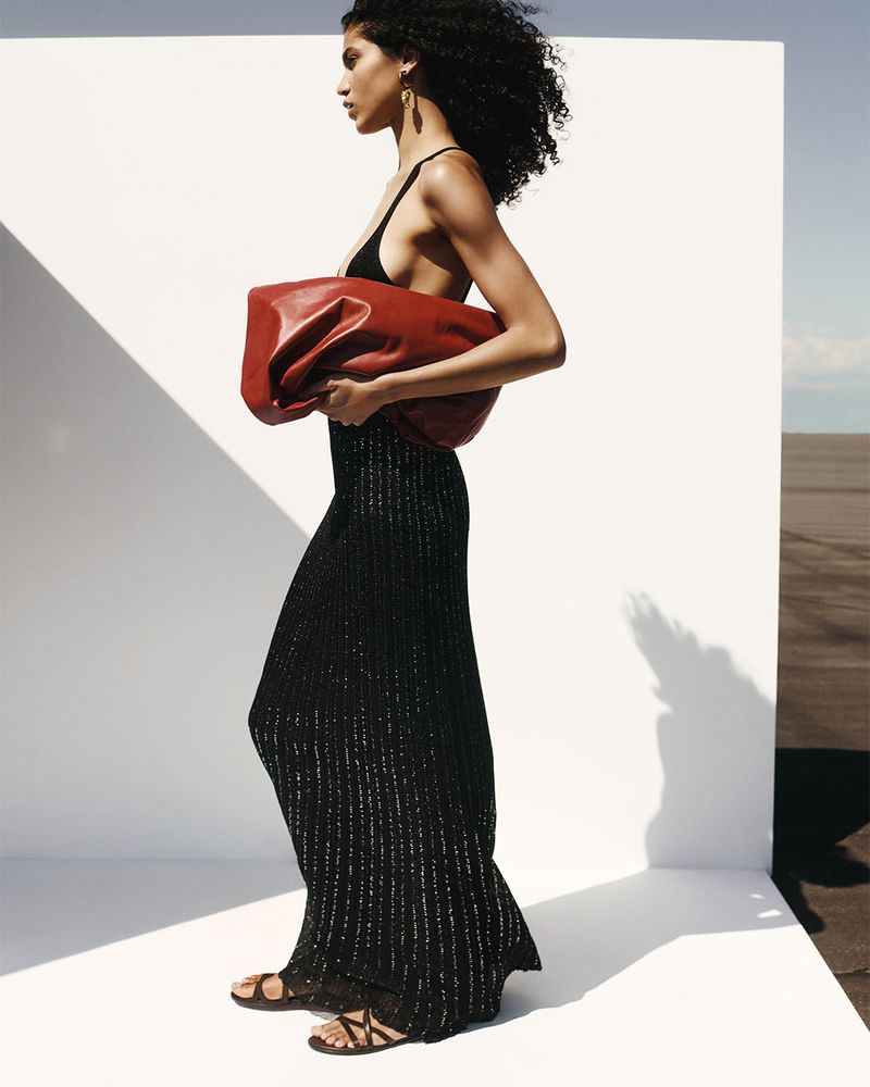 Fashion editorial shot of a Black woman carrying a red leather clutch, wearing a black dress and strappy sandals, against a white wall.