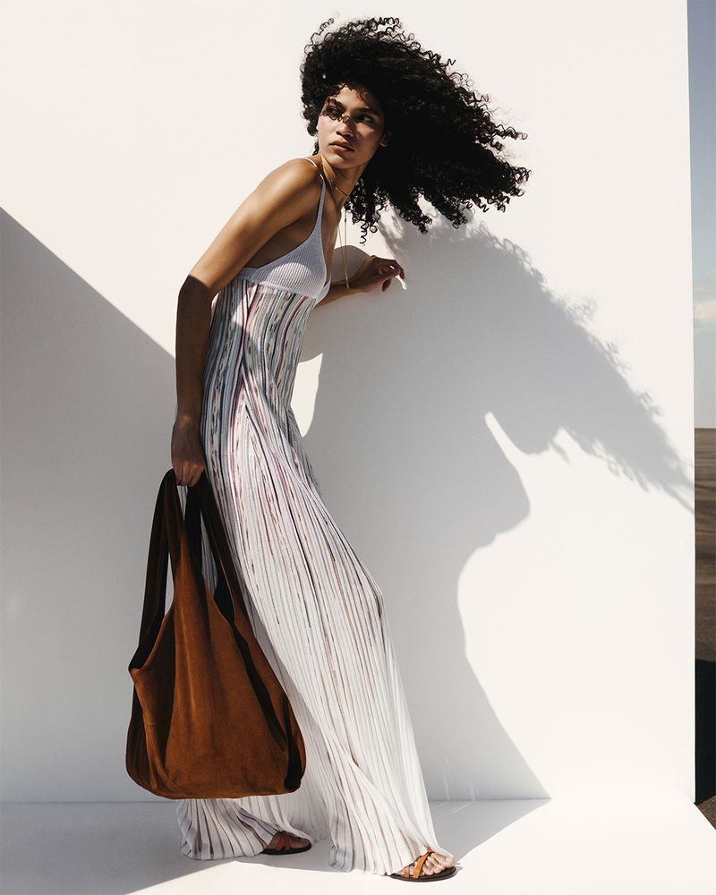 An adult female model with an afro poses in direct sunlight near a white wall wearing a maxi dress and sandals, and holding a brown bag.