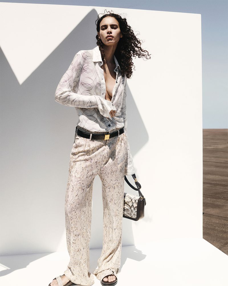 A woman in a patterned blouse and wide-leg pants poses against a white wall and dry terrain wearing sandals and carrying a snakeskin purse.