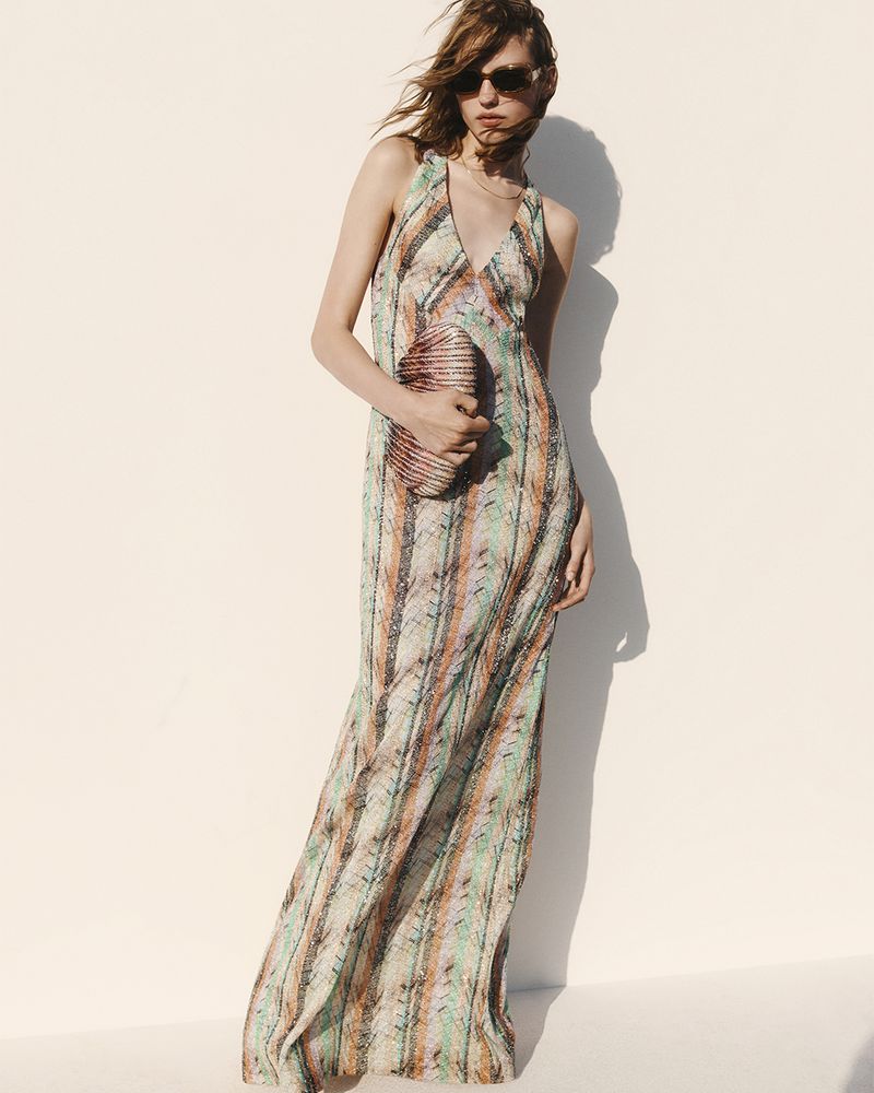 A woman poses in a long patterned halter dress and sunglasses, holding a beaded clutch, against a simple bright white background.
