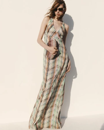A woman poses in a long patterned halter dress and sunglasses, holding a beaded clutch, against a simple bright white background.
