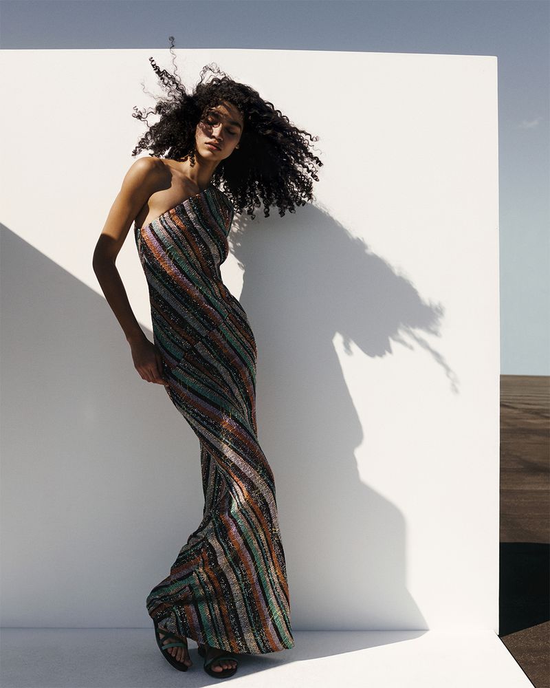 Elegant woman in a striped one-shoulder gown poses against a bright white wall under the sky. Fashion editorial photography.