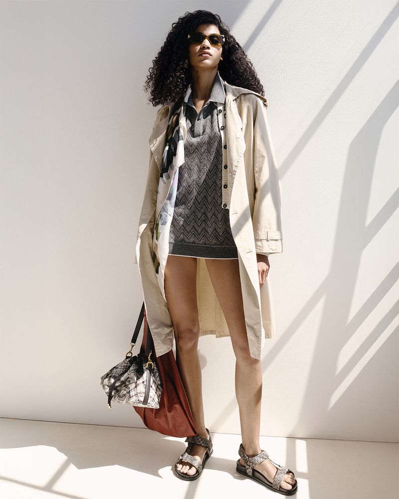 A woman with curly hair wearing a trench coat, knit polo dress, sandals, and carrying a tote bag poses in a brightly lit indoor space.