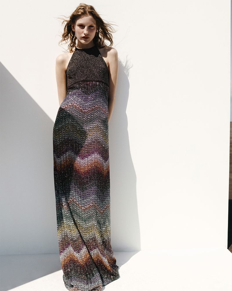 An adult female fashion model in a halter-style chevron-patterned long dress poses in front of a white wall in bright daylight.