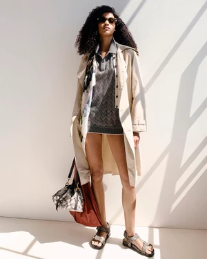 Model wearing sunglasses, a beige trench coat, and a grey knit mini dress holding bags against a sunlit white wall.