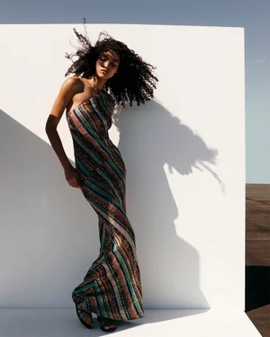 Model with dark curly hair wearing a striped one-shoulder dress against a white wall outdoors