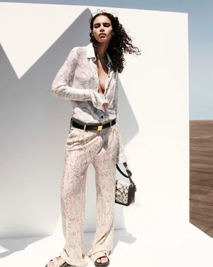 Model posing in a sheer patterned long-sleeve shirt and matching wide-leg trousers against white geometric panels in sunlight.