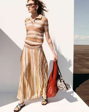 Model wearing a beige striped knit polo, matching pleated maxi skirt, eyeglasses, and holding a red leather tote bag in hard sunlight.