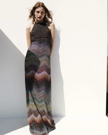 Model wearing a multicolored chevron knit halterneck maxi dress posing against a white backdrop in bright direct sunlight.