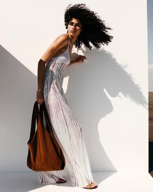Model with long dark curly hair wearing a white patterned long dress holding a large brown tote bag against a white wall.