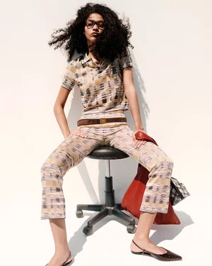 Model with dark curly hair wearing patterned polo and trousers seated on rolling stool against a white background.