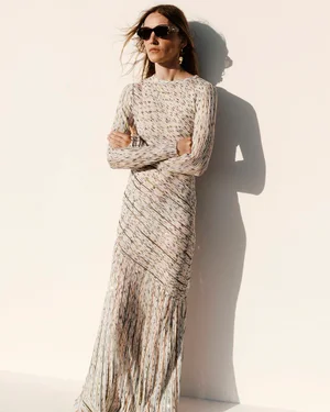 Model wearing dark sunglasses and a long-sleeve neutral knit maxi dress with fringe detailing against an off-white wall with sharp shadow.