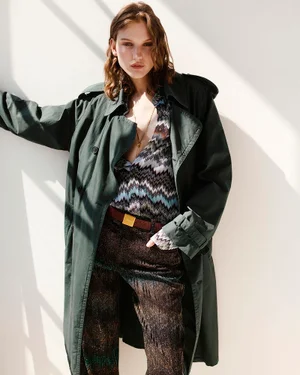 Model posing in an oversized dark green trench coat and patterned Missoni chevron shirt against a sunlit white wall.