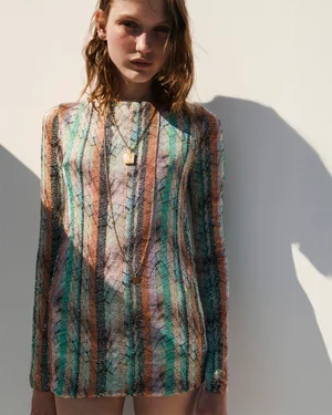 Model posing in a multicolored striped long-sleeve knit mini dress and gold pendant necklace against a sunlit wall.