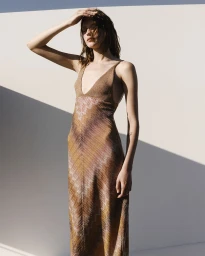 Model in a brown patterned slip dress posing outdoors against a stark white architectural wall with harsh diagonal shadows.