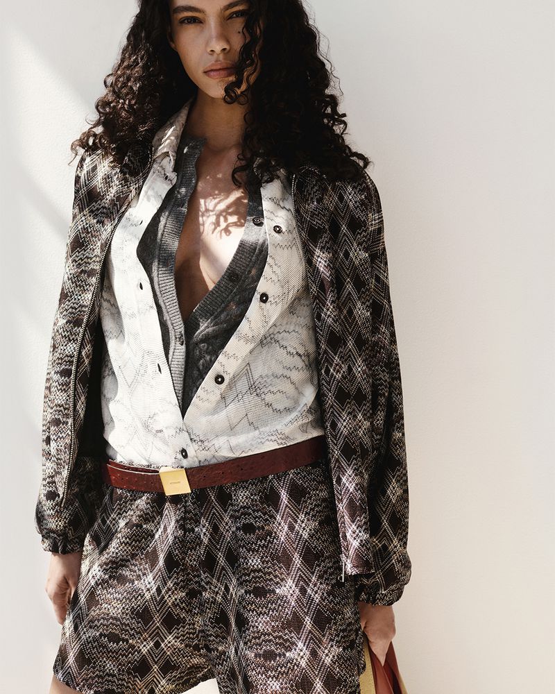 Model with dark curly hair wearing patterned jacket and shorts against a white background