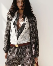Model with dark curly hair wearing patterned jacket and shorts against a white background