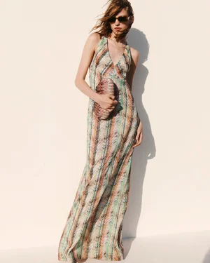 Model wearing a patterned multicolor halter maxi dress and dark sunglasses, holding a striped clutch bag against a white wall.