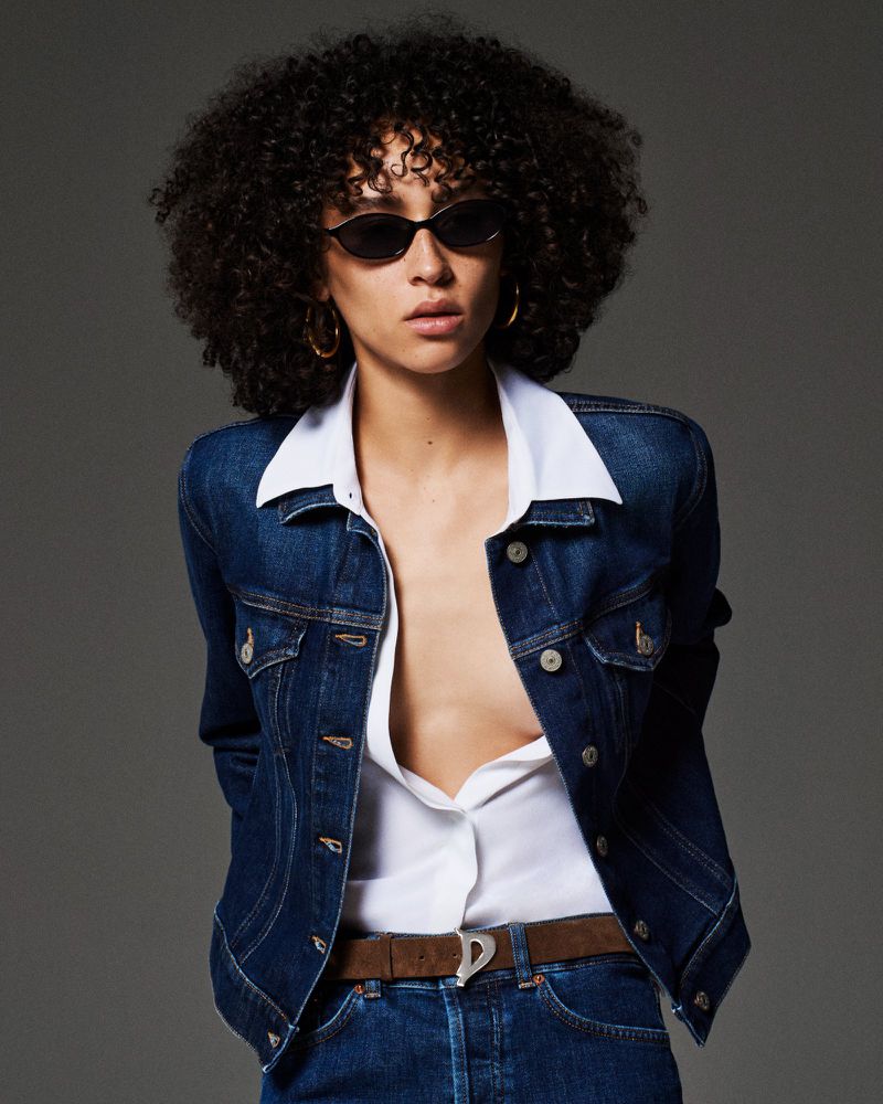 Model Lando van der Schee posing in a dark denim jacket, unbuttoned white shirt, and sunglasses against a grey studio backdrop.